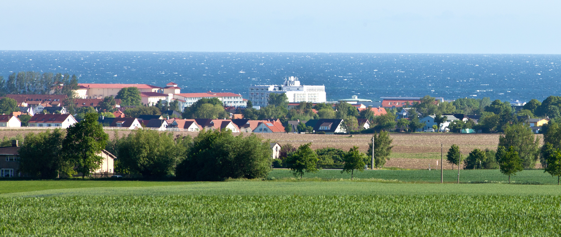 Panorama einer Küstenstadt mit roten Dächern, im Hintergrund das Meer und im Vordergrund Felder und Bäume.