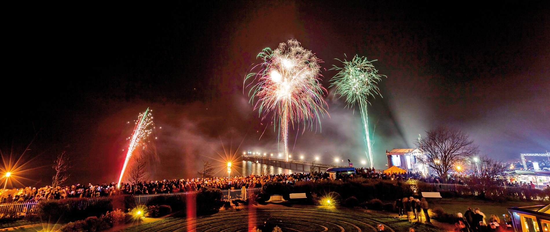 Menschenmenge beobachtet ein großes Feuerwerk in der Nacht, mit bunten Lichtern über einem Gewässer und einer beleuchteten Bühne.