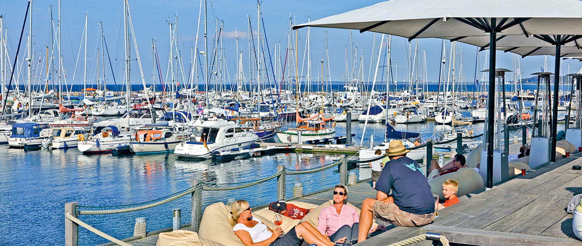 Ein Hafen mit vielen Segelbooten, im Vordergrund entspannen Menschen unter Sonnenschirmen.