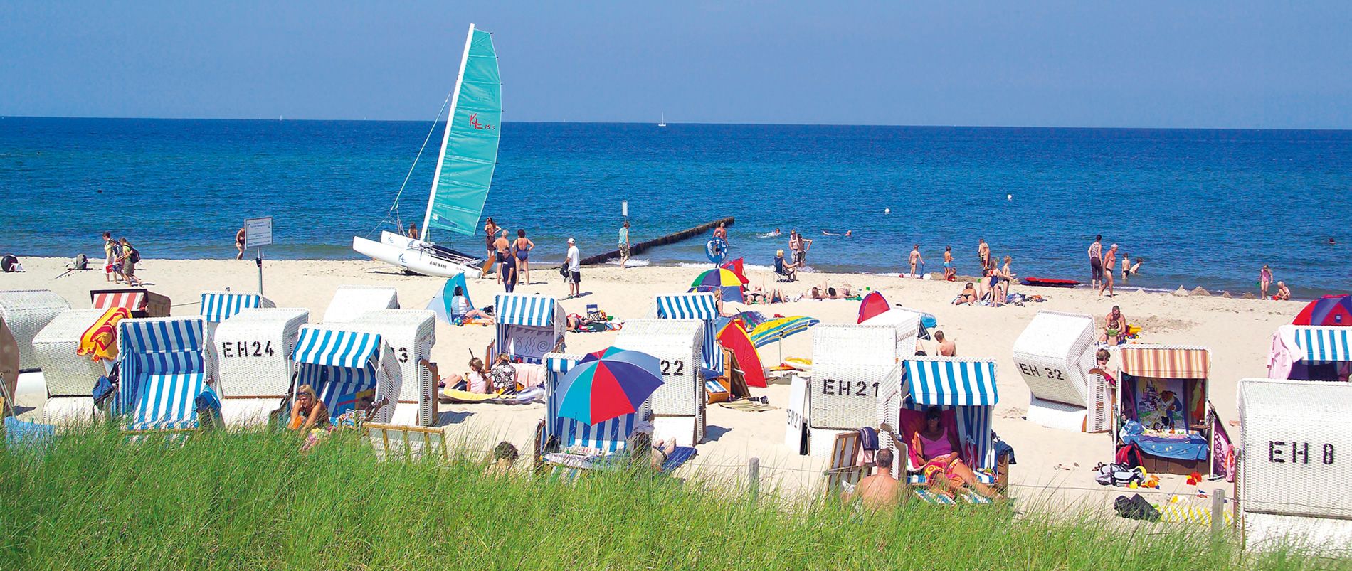 Strand mit bunten Strandkörben und einem Segelboot am Meer.