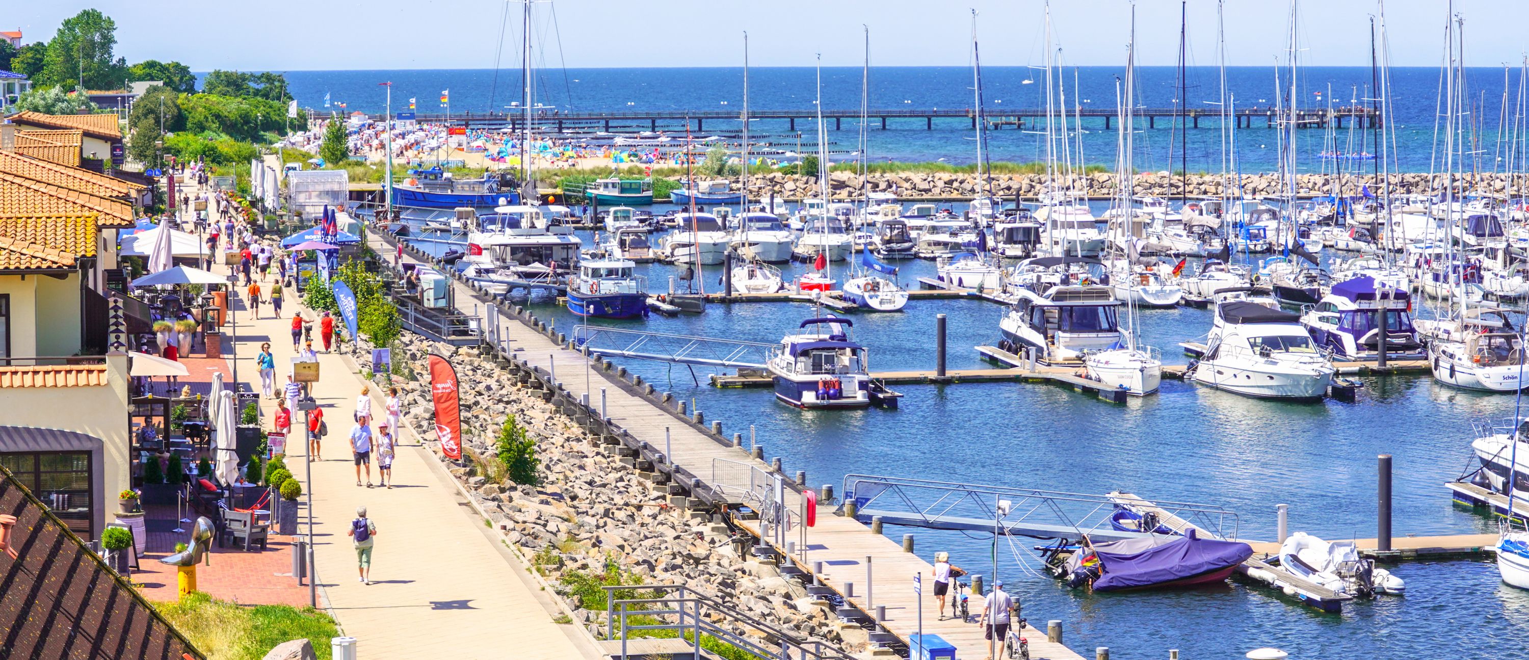 Ein Hafen mit vielen Segelbooten, im Vordergrund entspannen Menschen unter Sonnenschirmen.