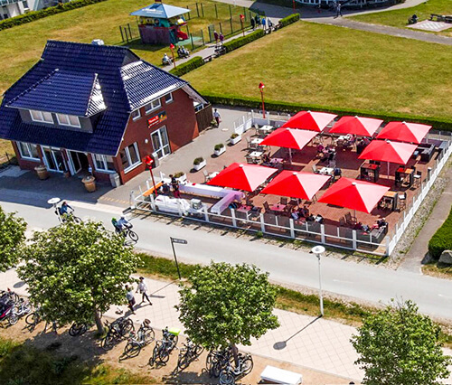 Luftaufnahme eines Gebäudes mit Terrasse und roten Sonnenschirmen, umgeben von einem Fahrradweg und Bäumen.