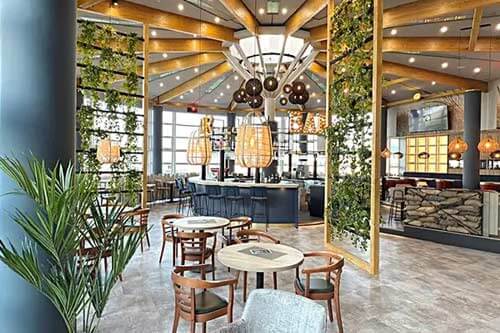 Modernes Restaurant mit offener Gestaltung, viel Natur und hängenden Pflanzen. Runde Holztische mit Stühlen, eine zentrale Bar und große Fenster sorgen für eine helle Atmosphäre.