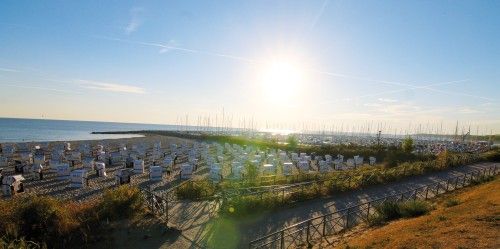 Sonnenuntergang über einem Strand mit zahlreichen Strandkörben und einem angrenzenden Hafen mit Segelbooten.