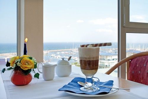 Gemütlicher Tisch mit Kaffee und Blumengesteck am Fenster mit Meerblick und Hafen im Hintergrund.