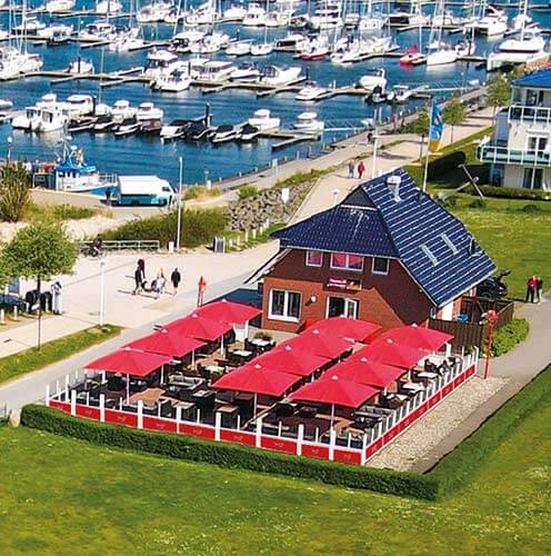 Luftaufnahme eines Hafenbereichs mit einem rot gedeckten Cafégarten und angelegten Booten.