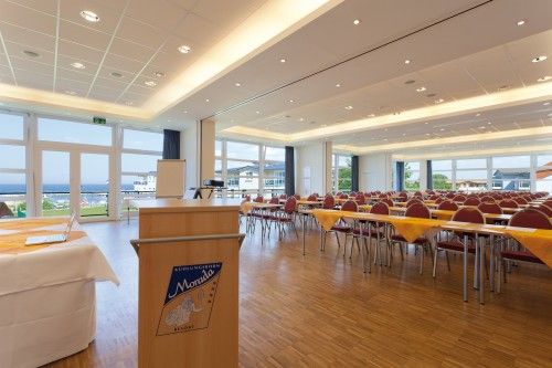 Heller Konferenzraum mit Rednerpult, Tischen und Stühlen, großen Fenstern mit Blick nach draußen.