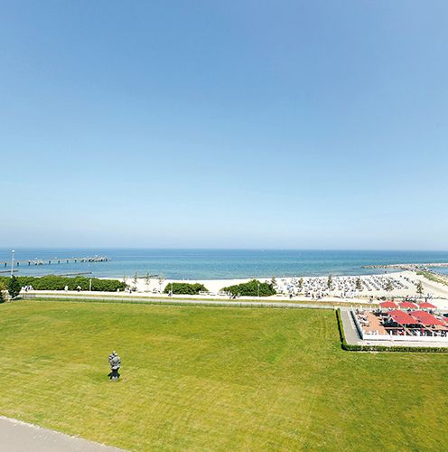 Blick auf eine Küstenlandschaft mit grünem Rasen, einem weißen Sandstrand und ruhigem, blauem Meer unter wolkenlosem Himmel.