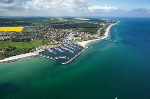 Luftaufnahme einer Küstenstadt mit Yachthafen und Sandstrand, umgeben von grünen Feldern und blauem Meer.
