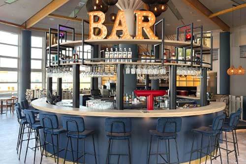 Moderne Bar mit kreisförmiger Theke, blauen Barhockern und beleuchtetem 'BAR'-Schriftzug.