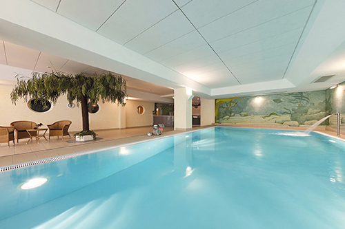 Innenpool mit Wandgemälde und Pflanzen, daneben Sitzbereich mit Stühlen.