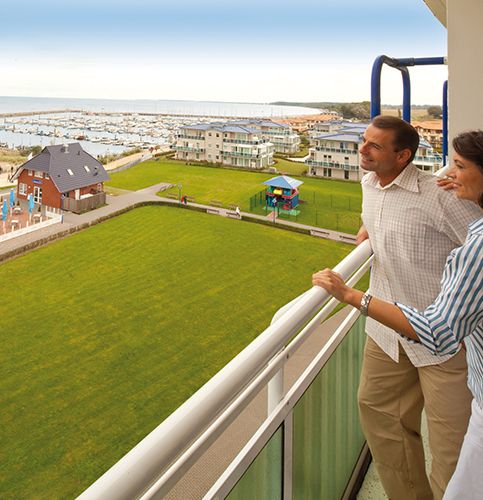 Ein Paar steht auf einem Balkon und blickt auf einen Yachthafen und moderne Gebäude.