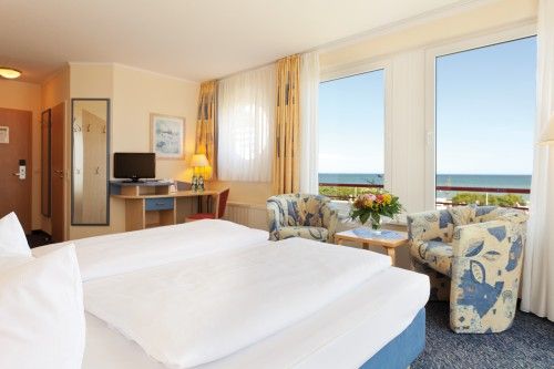 Helles Hotelzimmer mit einem Doppelbett, zwei Sesseln und einem Tisch mit Blumenschmuck. Große Fenster bieten einen Ausblick auf das Meer.
