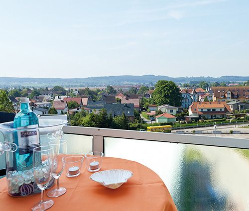 Blick von einem Balkon mit Tisch, dekoriert mit Gläsern und Flasche, auf eine ruhige Wohngegend mit mehreren Häusern im Hintergrund.
