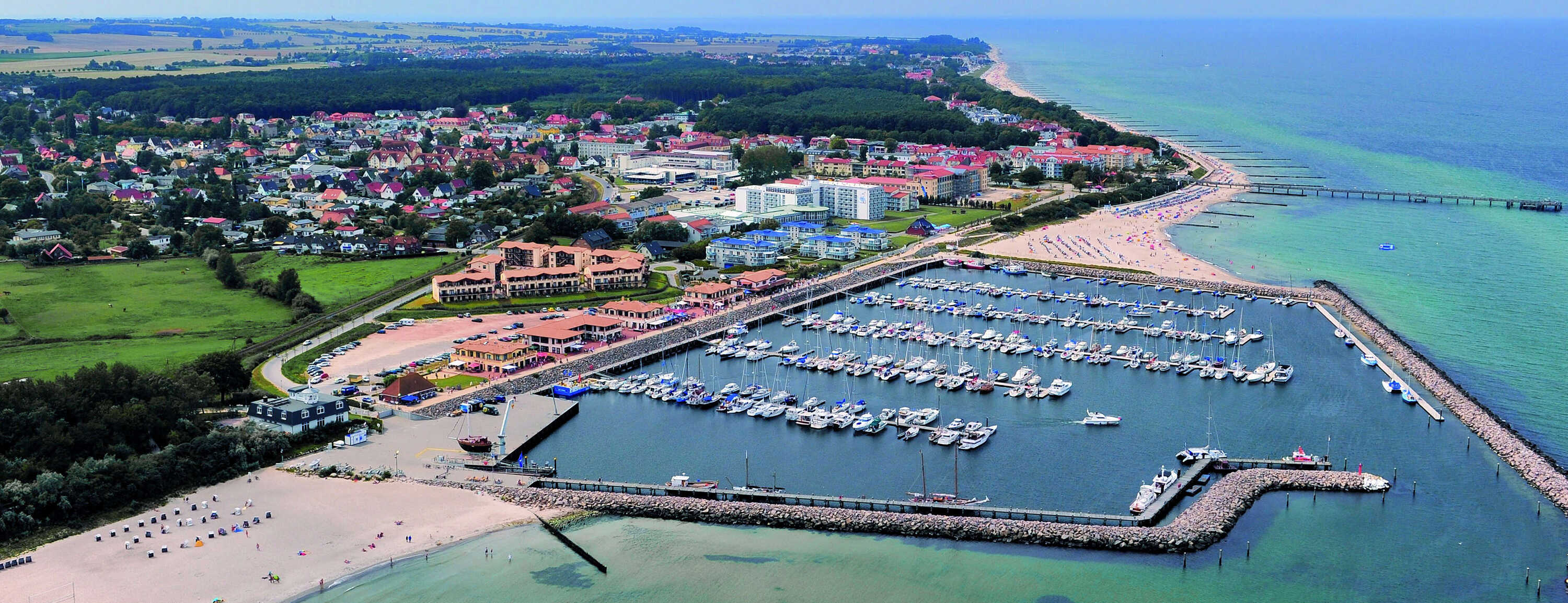 Luftaufnahme eines Hafens mit zahlreichen Booten, umgeben von einem Strand und einer Stadtlandschaft im Hintergrund.